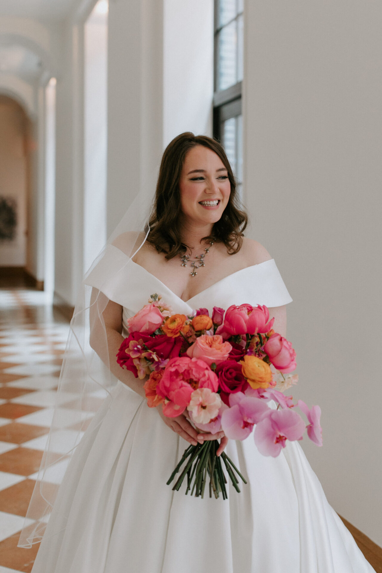 A bride in an off-the-shoulder white gown holds a large pink and orange bouquet while standing in a sunlit hallway with checkered floors.