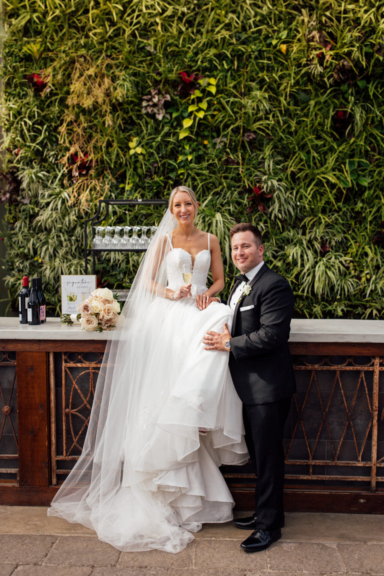 The bride is sitting on a countertop, and her groom is standing beside her in an outdoor setting.