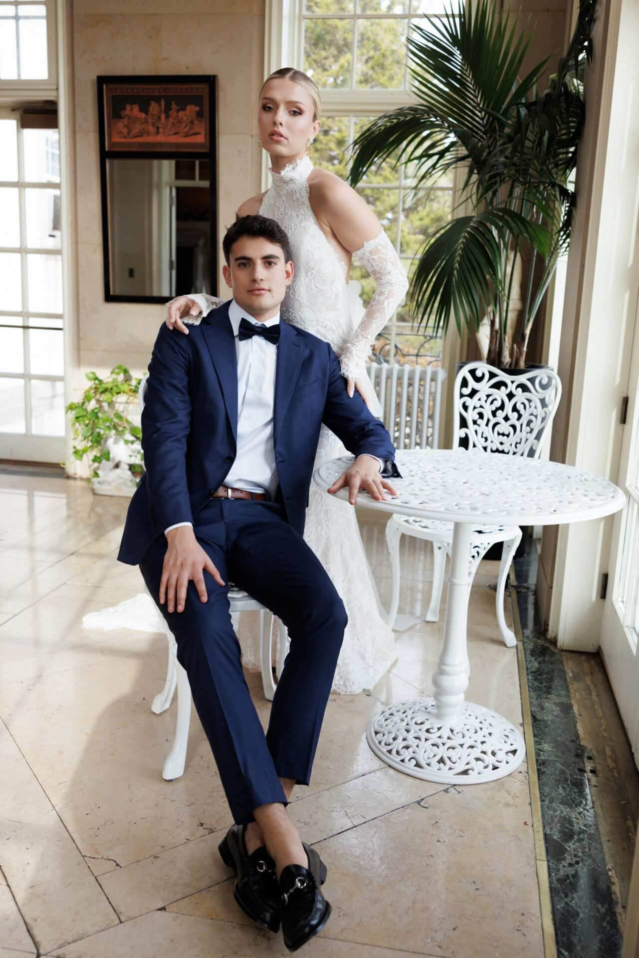 Groom in a navy suit sitting on a white chair beside a small garden table, posing confidently with his bride.