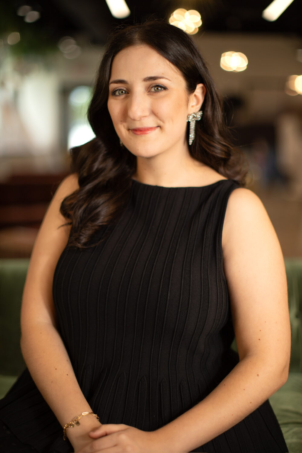 Smiling woman with long dark hair wearing a sleeveless black dress, seated against a softly blurred indoor background.