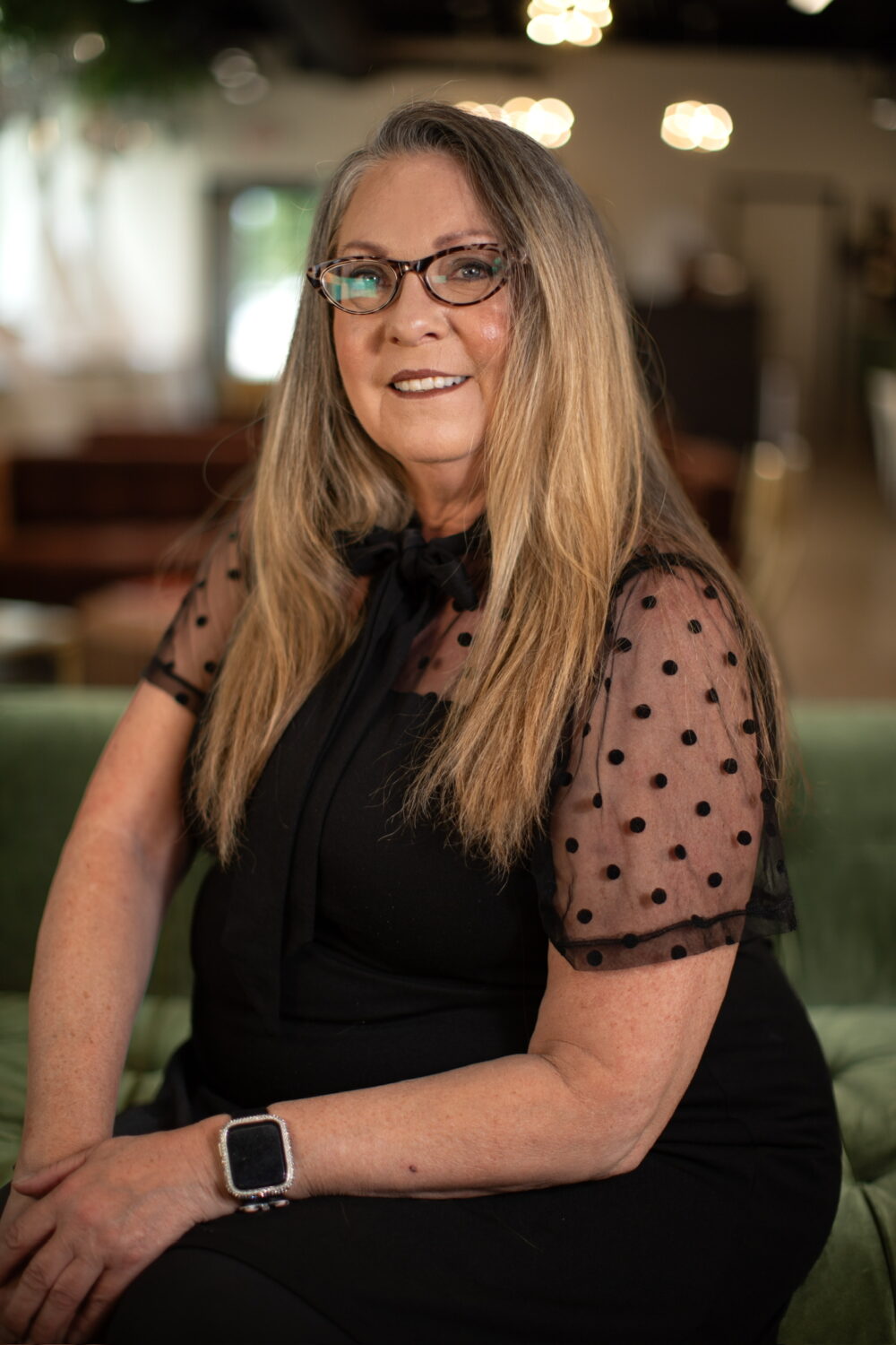 Smiling woman with long blonde hair and glasses wearing a black top with sheer polka-dot sleeves, seated in warm indoor lighting.