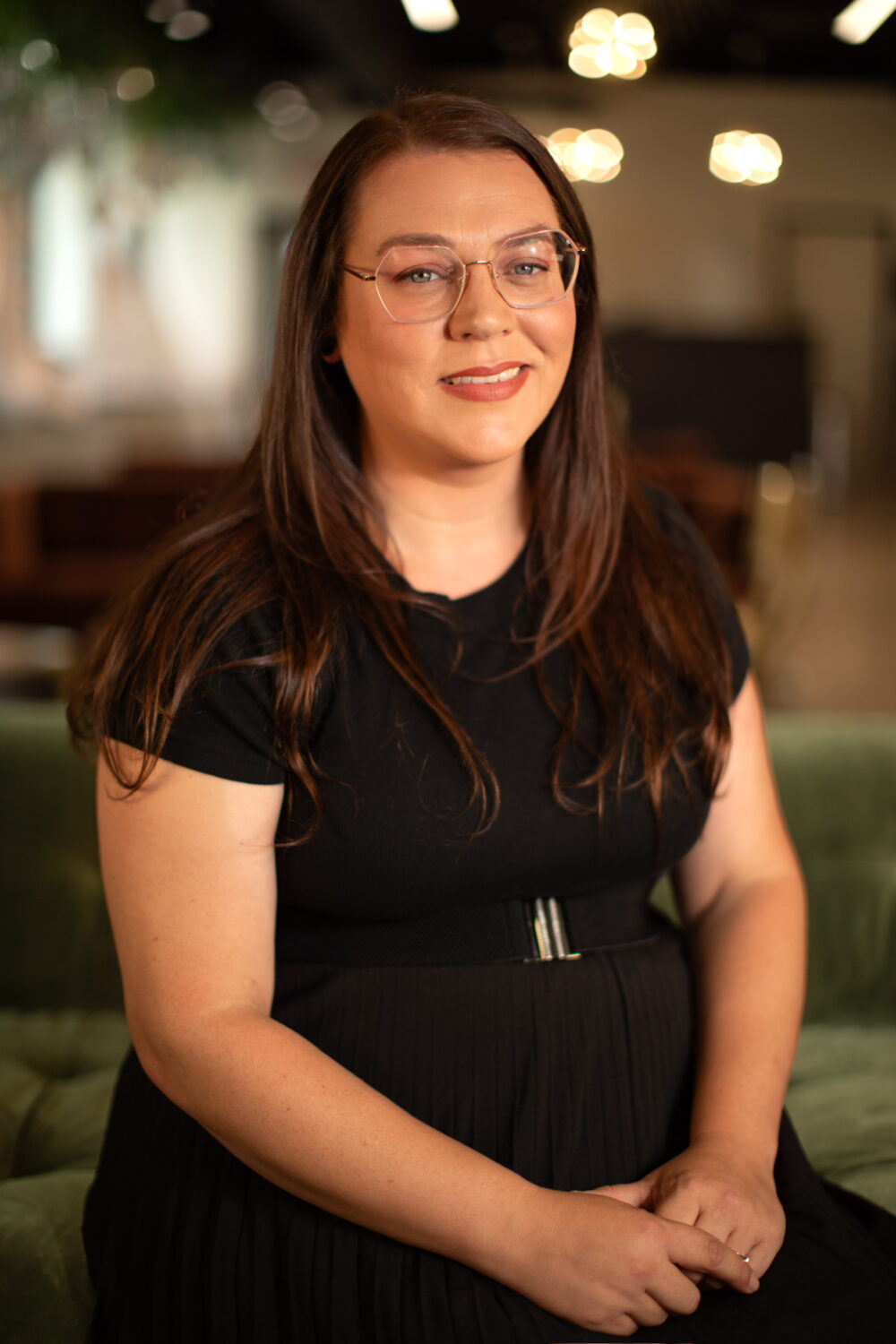 Smiling woman with long brown hair and glasses wearing a black dress, seated in a softly lit indoor environment.