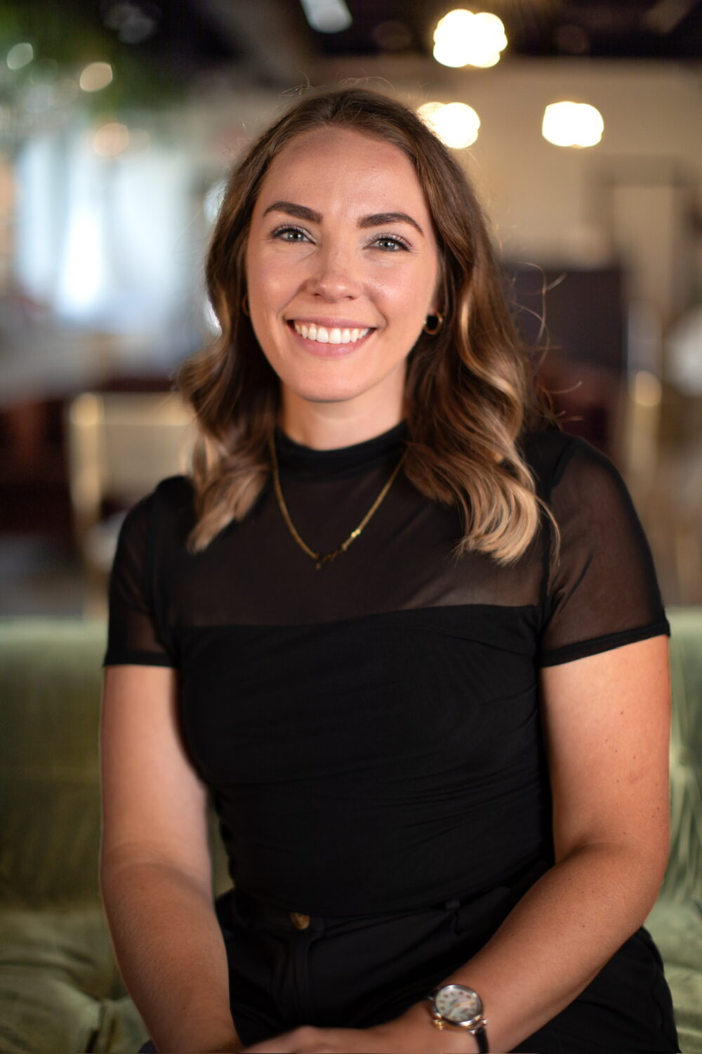 Smiling woman with wavy light brown hair wearing a black short-sleeve dress, seated against a warm, softly blurred indoor background.