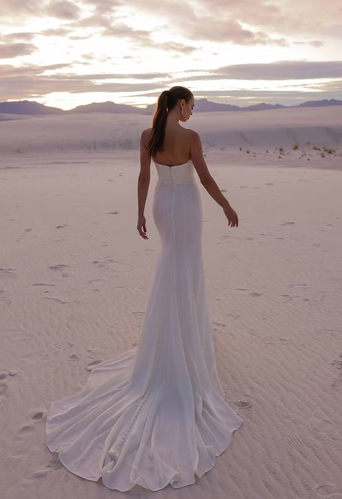 Back view of a bride walking across white sand dunes in a fitted white gown with a long train under a cloudy sky.