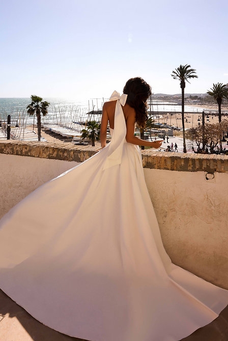 A bride in a flowing white gown viewed from behind, leaning on a terrace wall with a coastal marina and palm trees in the distance.