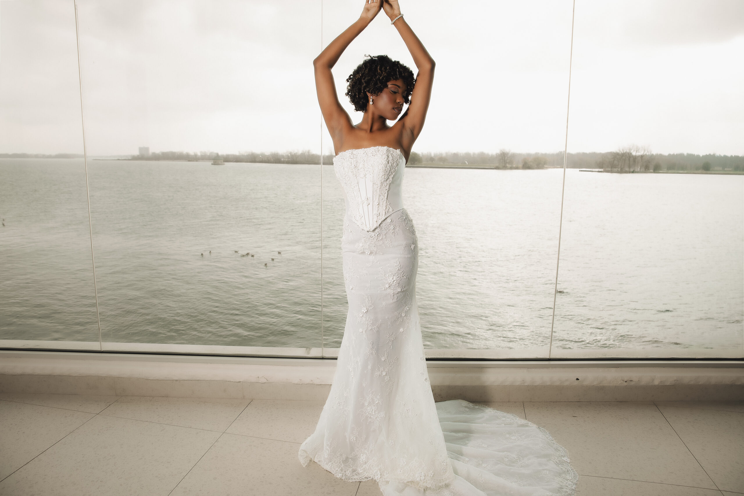 A bride in a fitted white gown poses indoors near large windows, raising her arms above her head.