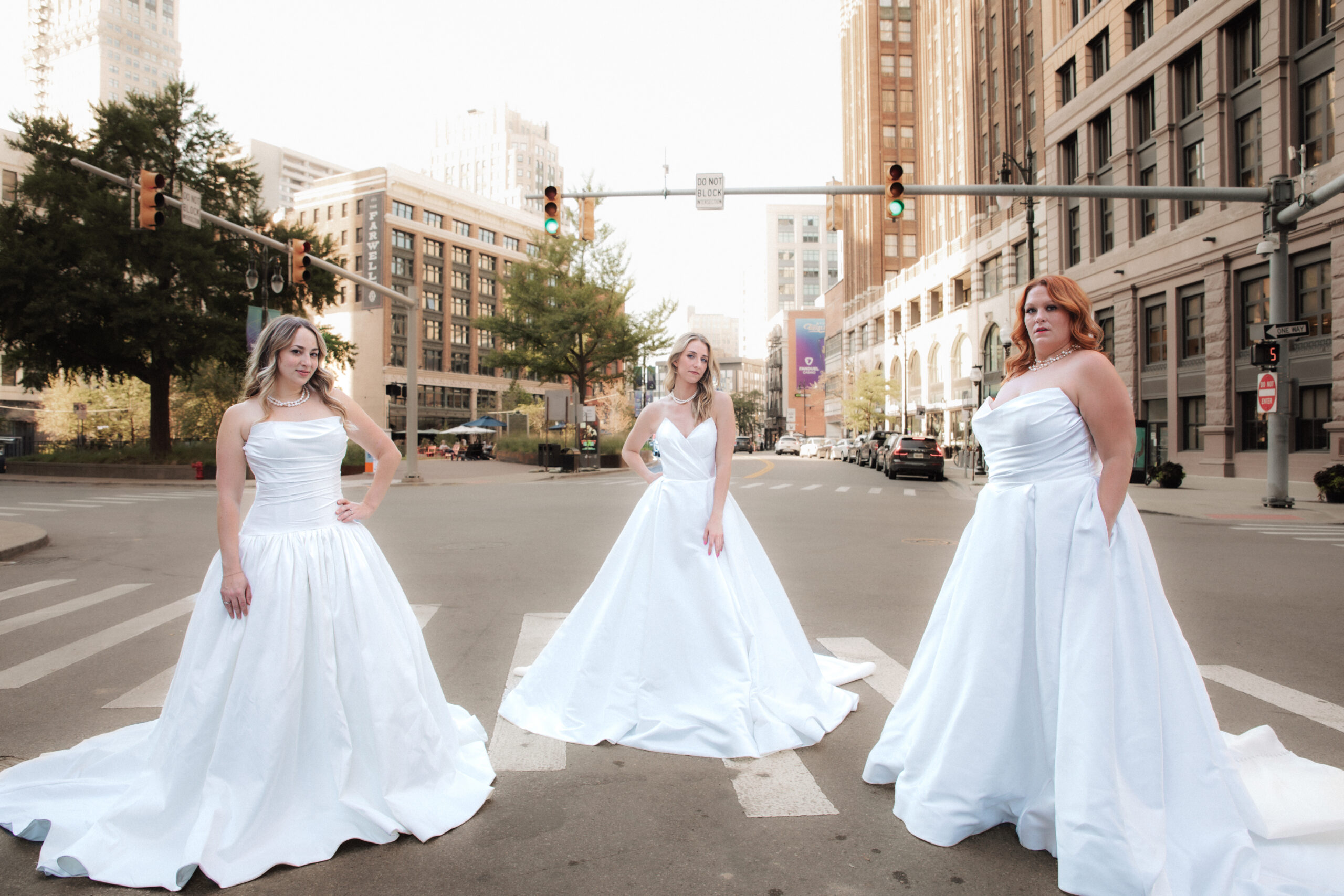 Three brides in flowing white dresses pose together across a city street, holding their gowns.