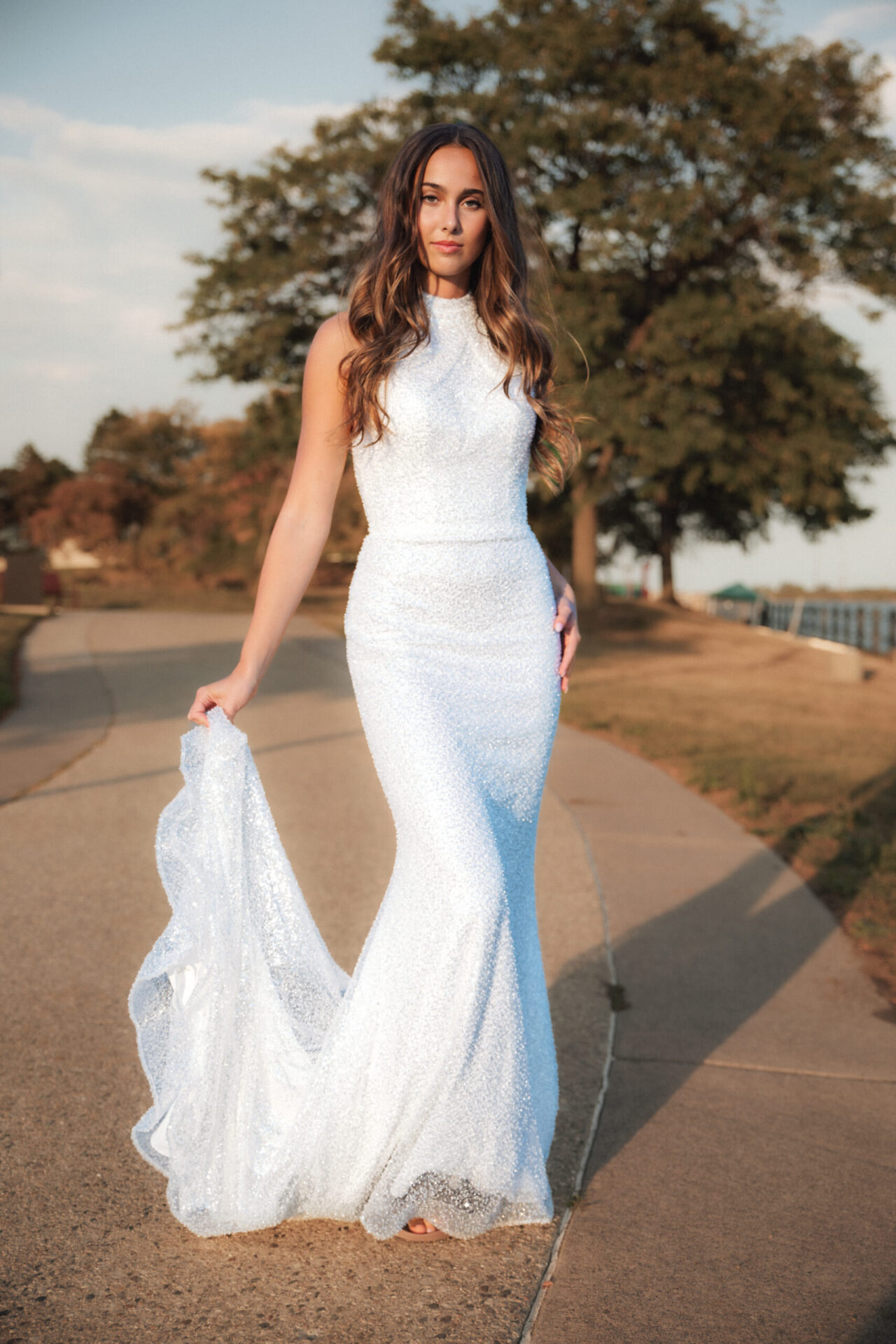 A bride in a sleek white gown walks along a paved path outdoors, holding part of her dress.