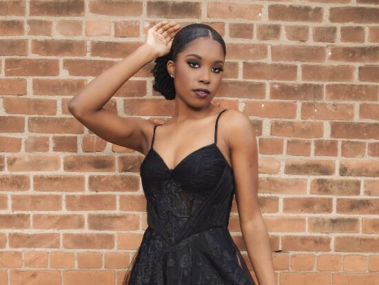 A woman wearing a black lace gown with thin straps poses in front of a brick wall with one hand raised near her head.