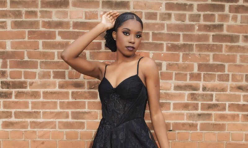 A woman wearing a black lace gown with thin straps poses in front of a brick wall with one hand raised near her head.