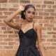 A woman wearing a black lace gown with thin straps poses in front of a brick wall with one hand raised near her head.