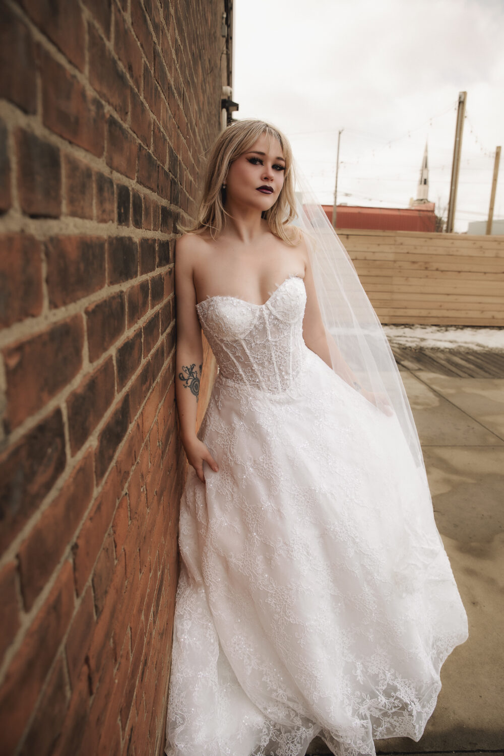 A bride in a strapless lace wedding dress and veil leans against a brick wall outdoors on a wooden deck.