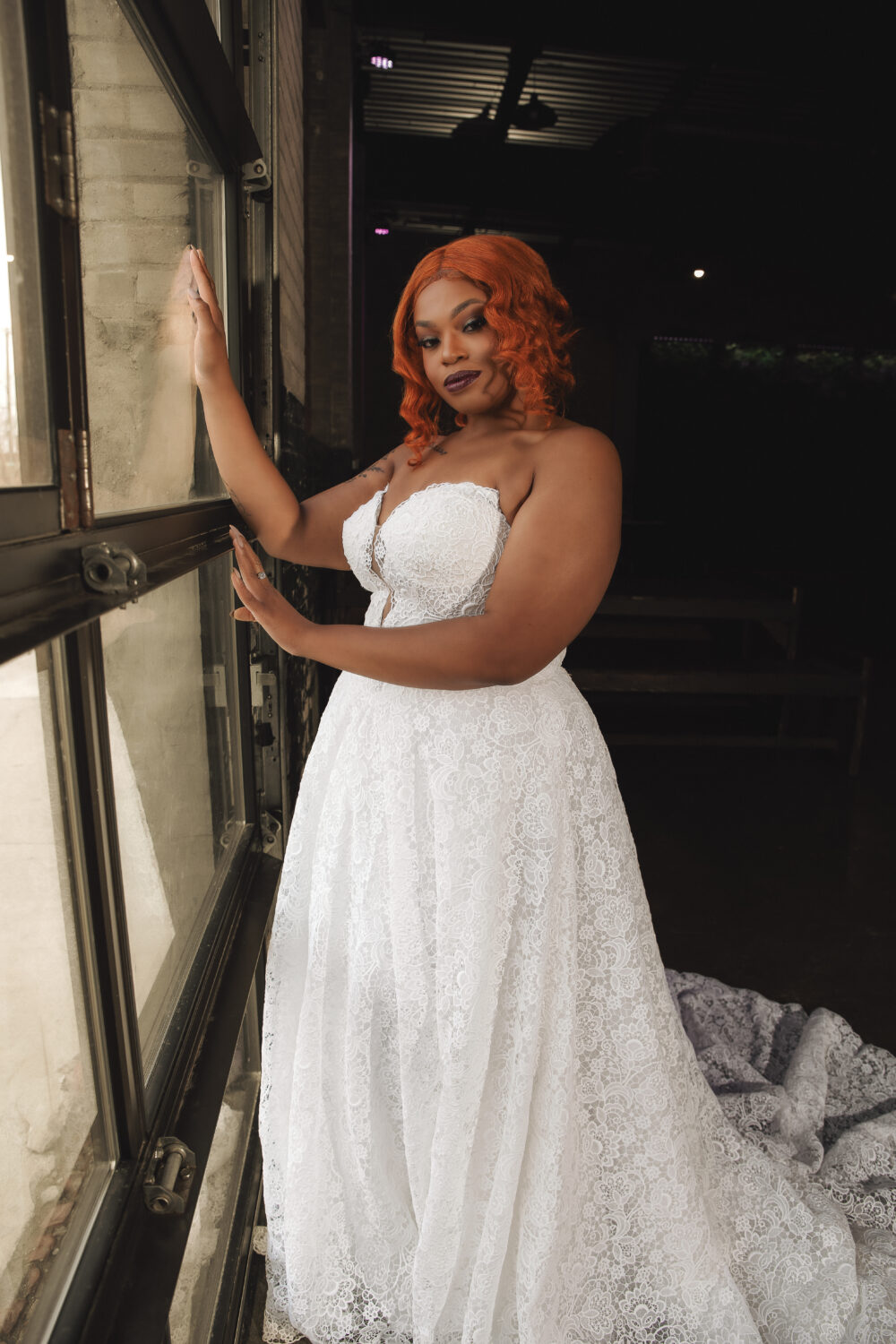 A bride with short red hair stands by a window wearing a strapless lace wedding gown with a long train.