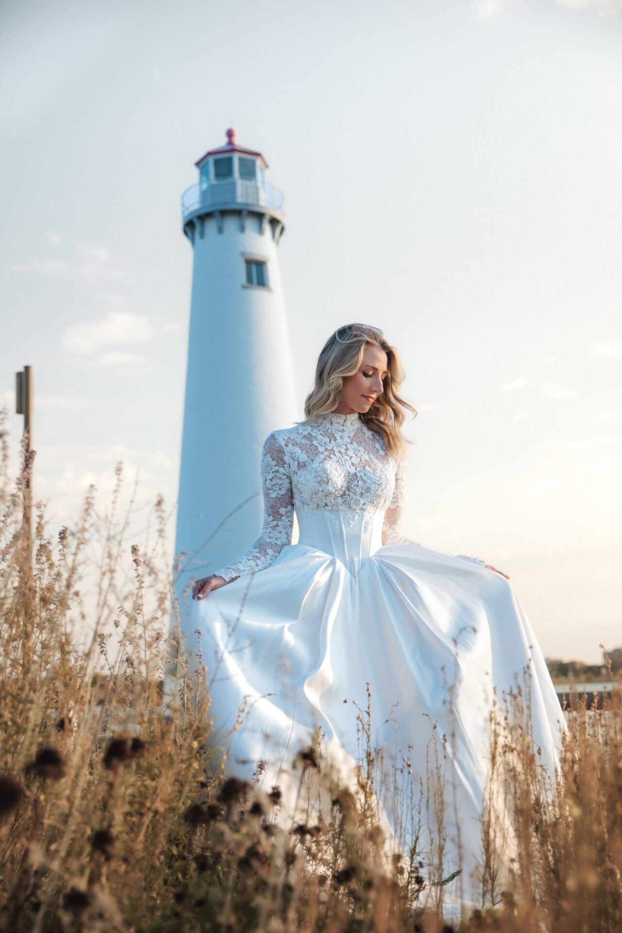 A woman in a light-colored dress sits near tall grass with a lighthouse in the background.