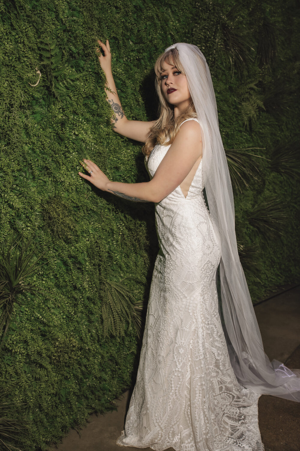 A bride in a fitted lace wedding dress with a long veil stands beside a green plant wall, gently touching the foliage.
