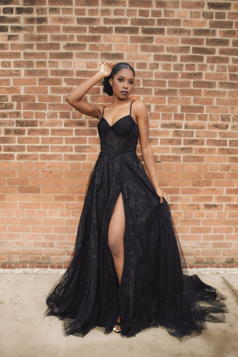 A woman wearing a black gown with a thigh-high slit poses against a brick wall while holding part of the skirt.