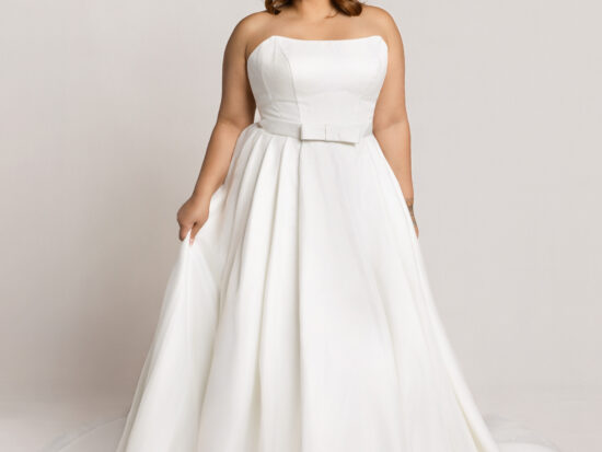 Smiling bride wearing a strapless white wedding gown with a fitted bodice and full skirt, holding the sides of the dress against a neutral background.