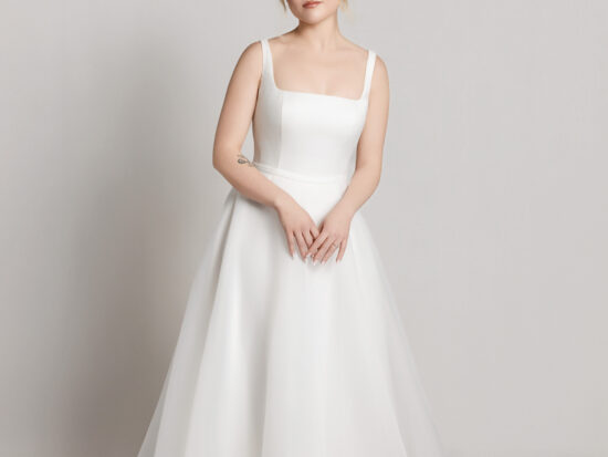 Bride wearing a soft tulle wedding dress with thin straps and a full skirt, standing with hands gently clasped.