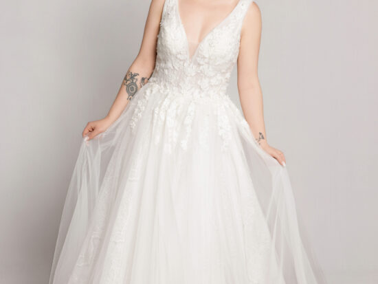 Bride wearing a sleeveless V-neck lace wedding gown with a full tulle skirt, holding the sides of the dress while standing against a neutral background.