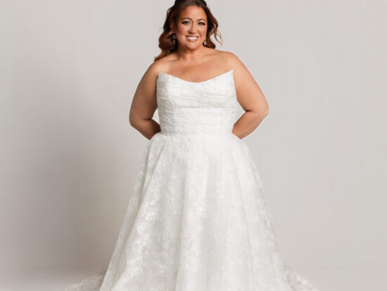 Bride wearing a one-shoulder lace wedding gown with a full skirt and train, standing with hands behind her back against a neutral background.
