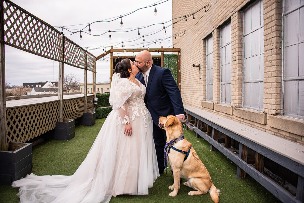 Newlywed couple kisses on a rooftop patio decorated with string lights while their dog sits beside them.