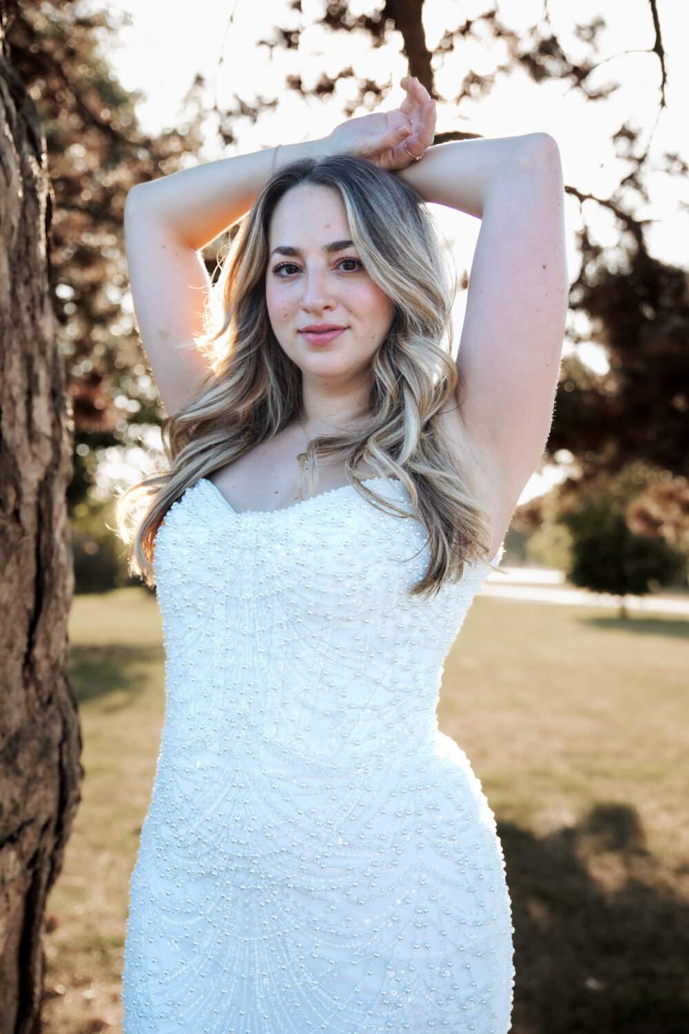 A close-up portrait of a bride in a strapless beaded wedding gown posing with her arms raised near a tree in a sunlit outdoor setting.