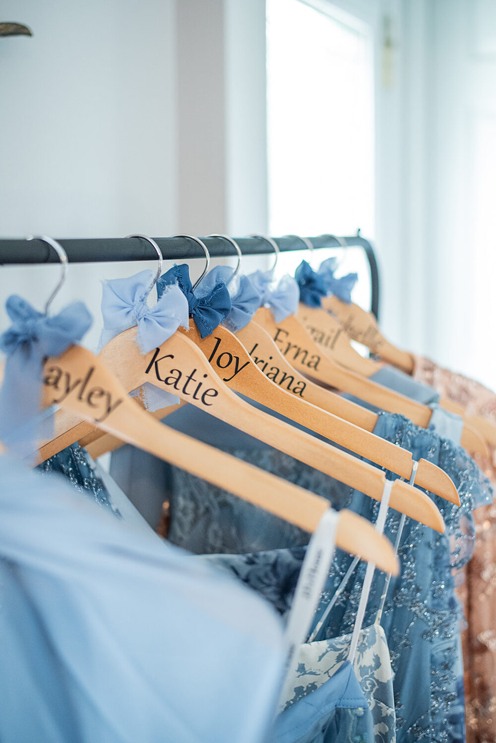 Bridesmaid dresses hanging on wooden hangers labeled with names and decorated with blue bows on a clothing rack.