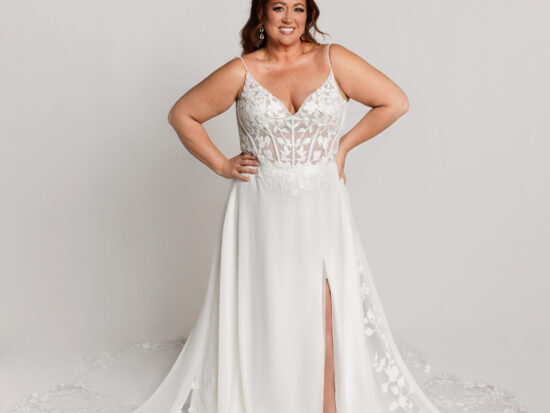 Smiling bride wearing a lace wedding gown with a fitted bodice, thin straps, and a high slit, standing with hands on hips.