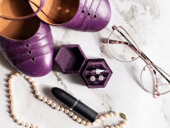 Wedding accessories flat lay with purple shoes, pearl necklace, lipstick, glasses, and ring box on a marble surface.