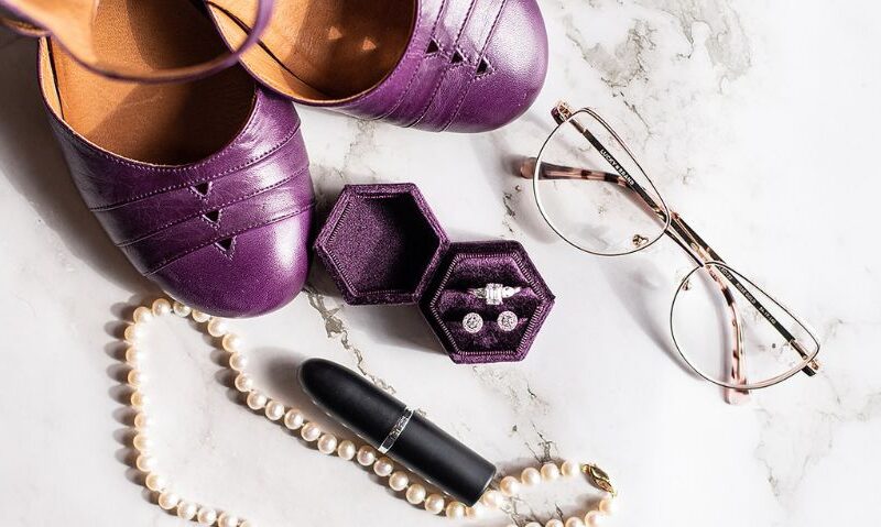 Wedding accessories flat lay with purple shoes, pearl necklace, lipstick, glasses, and ring box on a marble surface.