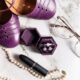 Wedding accessories flat lay with purple shoes, pearl necklace, lipstick, glasses, and ring box on a marble surface.