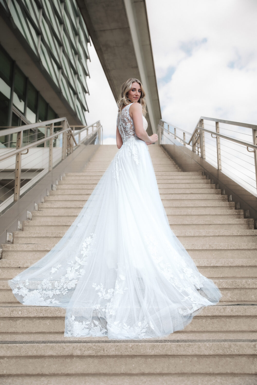 Bride stands on wide outdoor stairs in a voluminous gown with a long train, framed by modern architectural structures.
