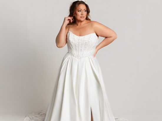 Bride wearing a strapless white gown with a front slit and long train, posing with one hand near her hair against a neutral background.