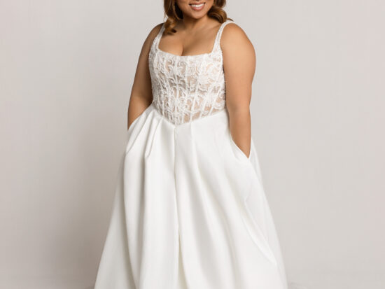 Smiling bride wearing a white wedding gown with a structured bodice and full skirt, standing with hands in pockets.
