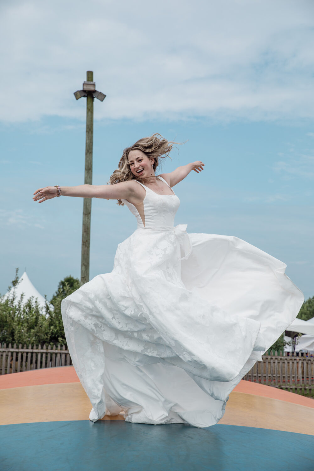 Bride twirls joyfully outdoors in a flowing wedding gown on a colorful circular platform under a cloudy sky.