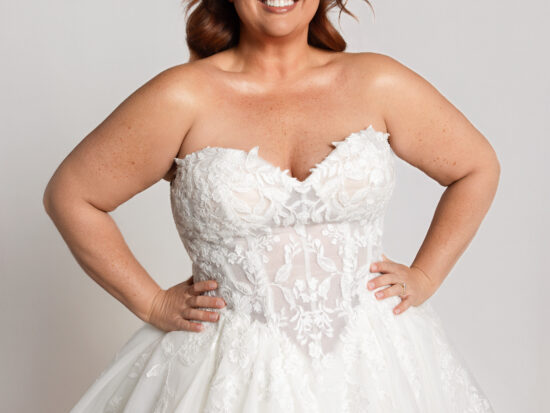 Smiling bride wearing a strapless lace wedding gown with a sweetheart neckline and full skirt, standing with hands on hips.