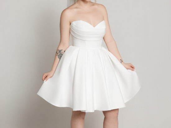 Bride wearing a short strapless white dress with a flared skirt, holding the hem slightly while standing against a neutral background.