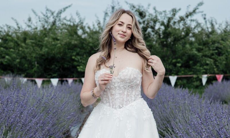 A bride in a strapless wedding gown stands between rows of purple flowers while holding a small stem and smiling toward the camera.