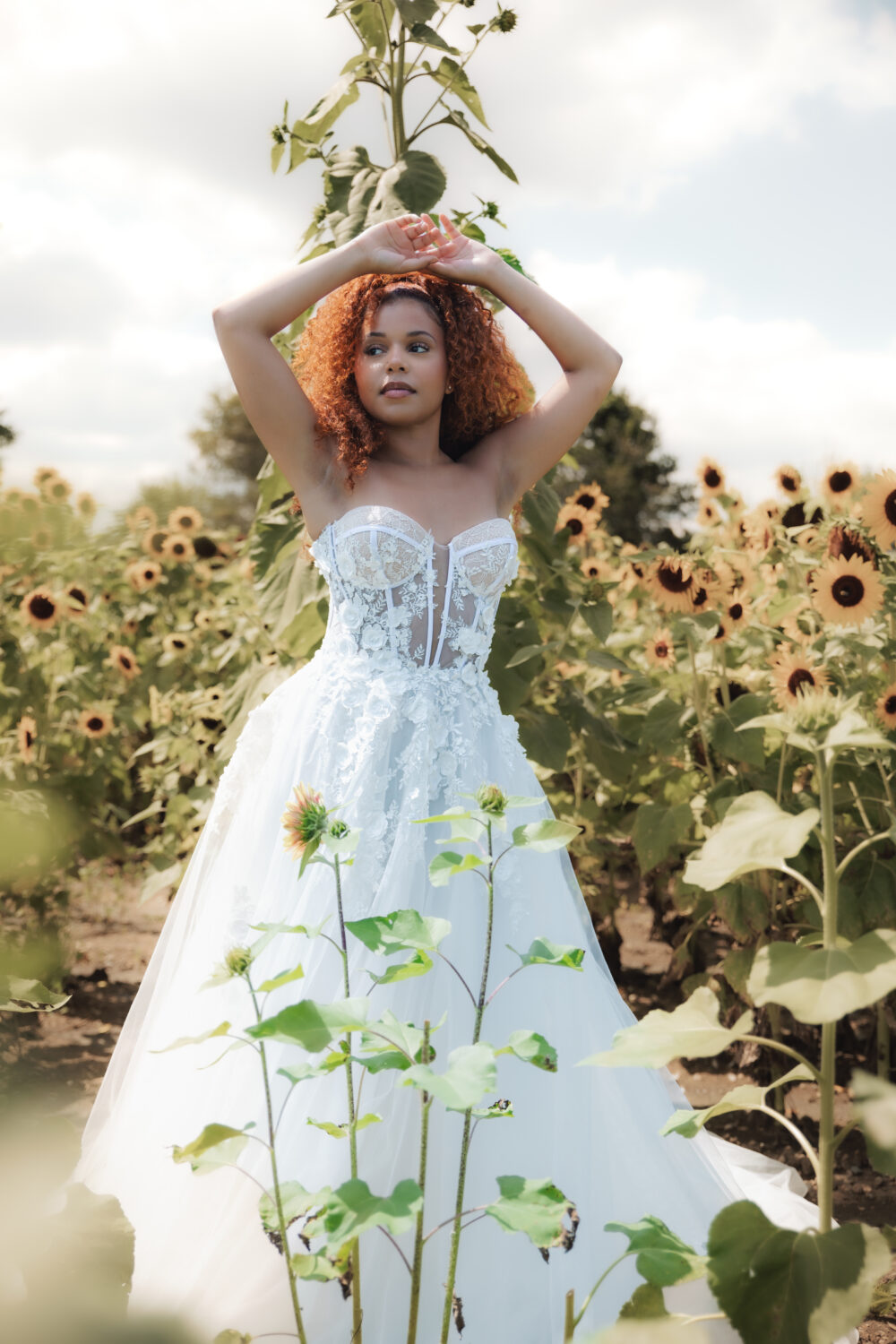 Close-up of bride in a lace bodice gown standing in a sunflower field with greenery and blooms surrounding her.
