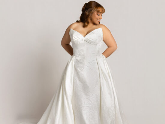 Bride wearing a strapless white wedding gown with a sweetheart neckline and full skirt, posing with hands in pockets and looking to the side.