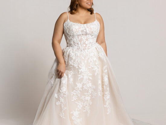 Bride wearing a lace wedding gown with thin straps and a full tulle skirt, holding the sides of the dress while standing against a neutral background.