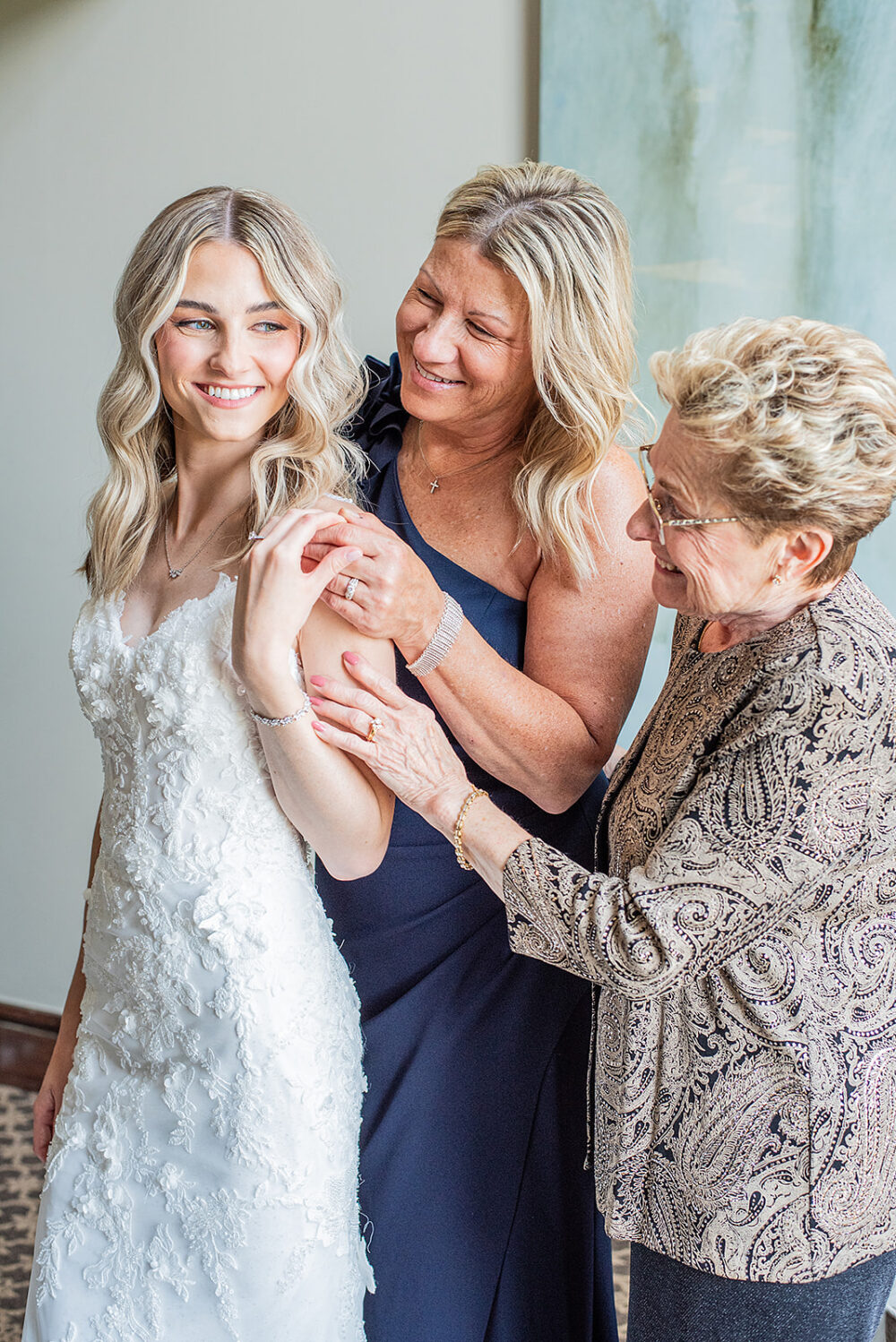 Bride smiling while two women help fasten jewelry and adjust her wedding dress.