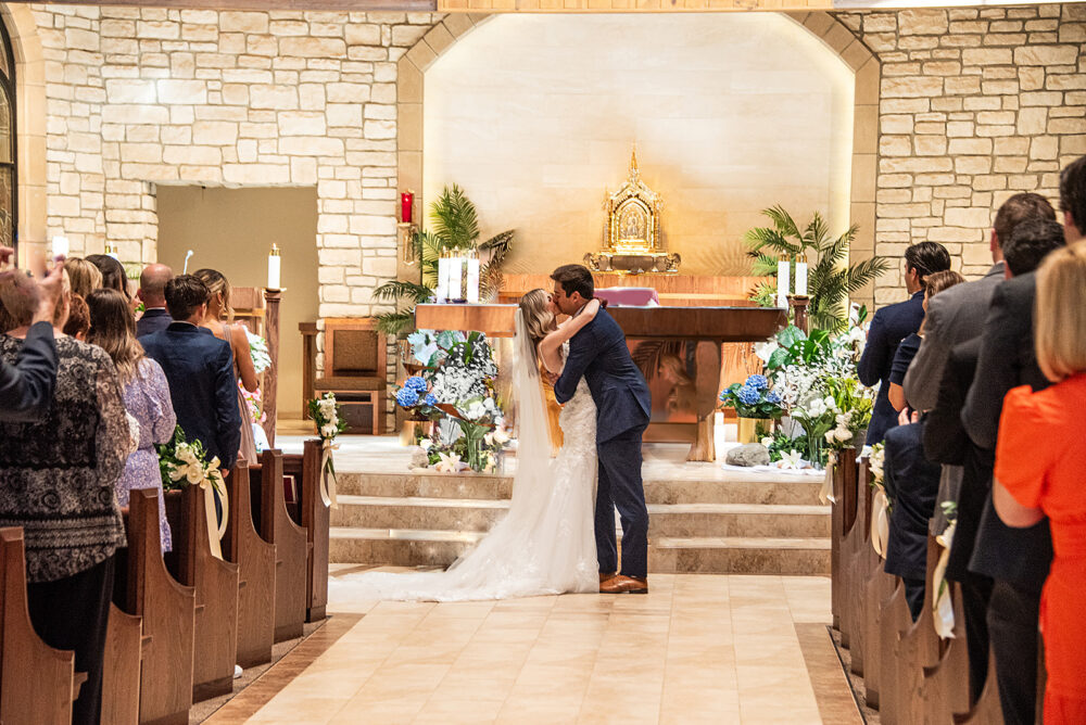 Bride and groom kiss at the altar during a church wedding ceremony while guests stand and watch.