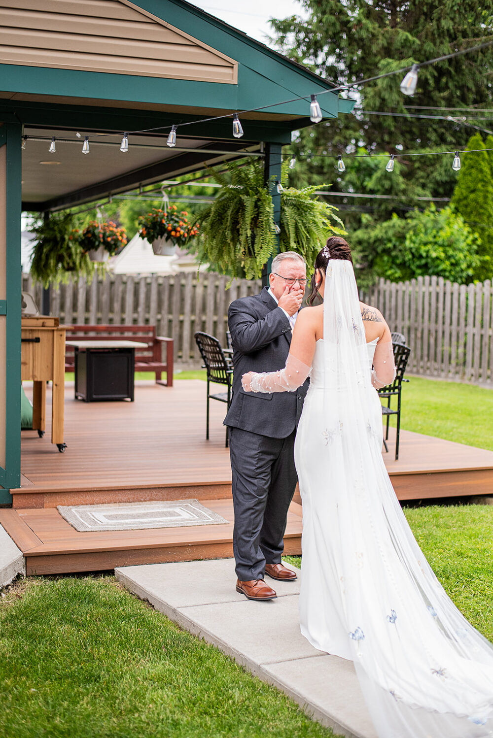 Bride shares an emotional first look with an older man on a backyard deck decorated with hanging plants and string lights.