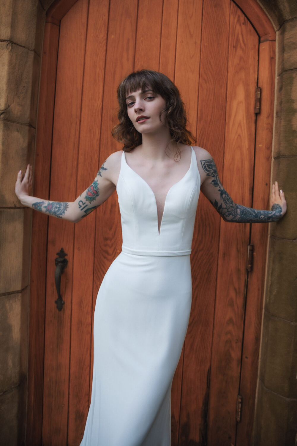 Woman in a sleek, fitted wedding dress with a deep neckline poses against a large wooden door with stone framing.