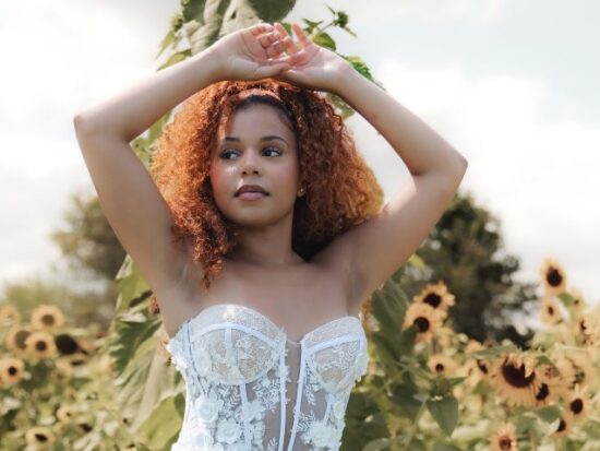 Bride in a lace wedding dress stands in a sunflower field with arms raised, surrounded by tall sunflowers in warm sunlight.
