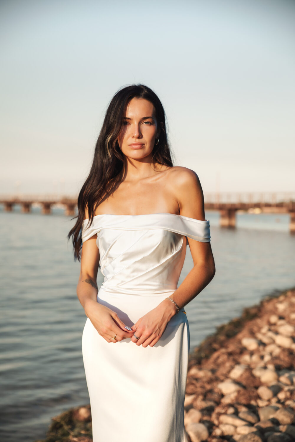 A bride in an off-the-shoulder satin wedding dress stands by the water with a bridge in the background during golden hour.