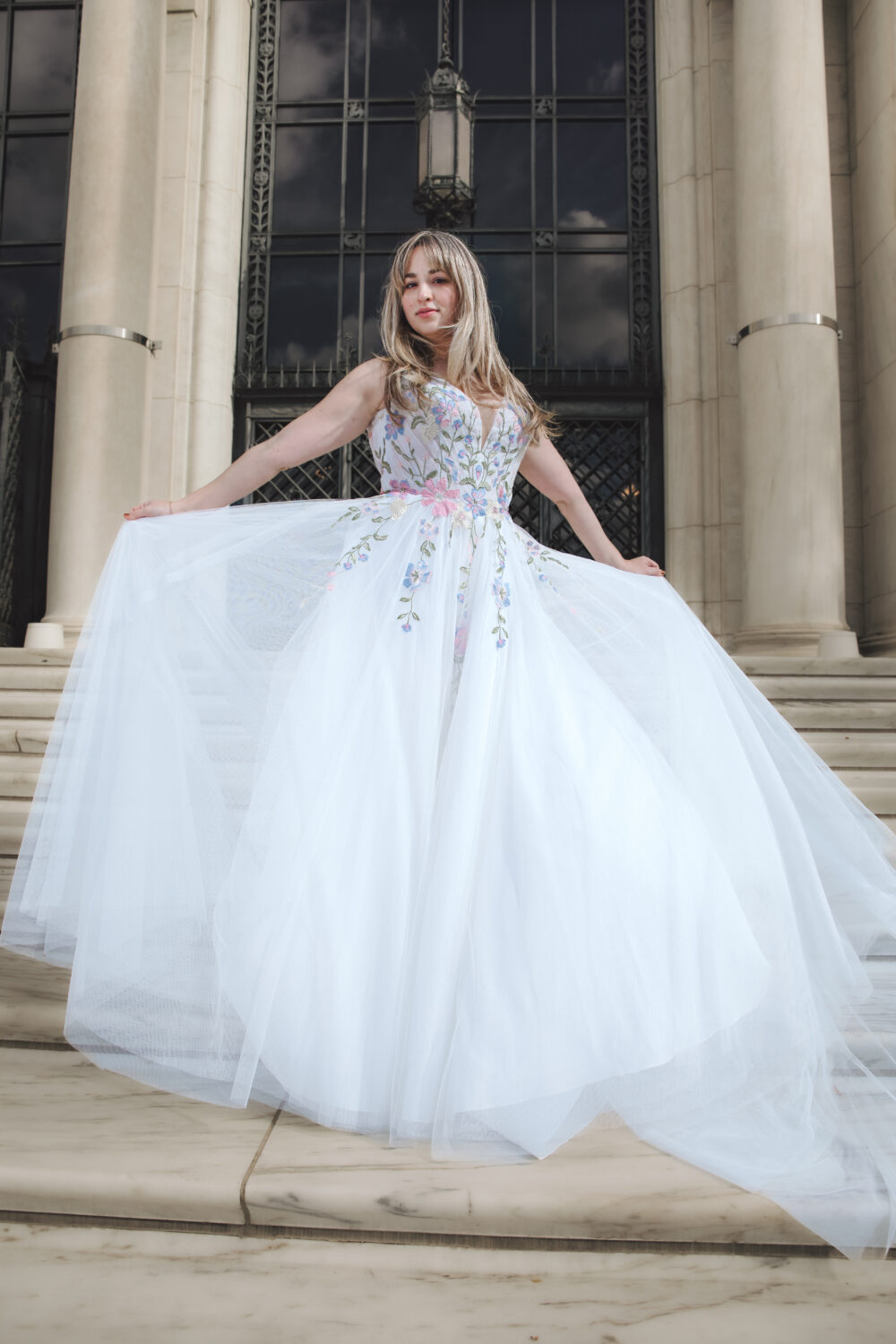 A bride in a voluminous ball gown with colorful floral embroidery holds out her skirt while standing on marble steps in front of a grand building.