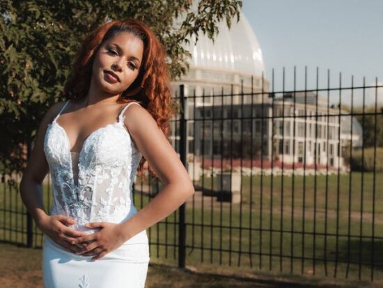 A bride with red hair wears a fitted lace wedding dress while standing outdoors near a glass conservatory behind a black fence.