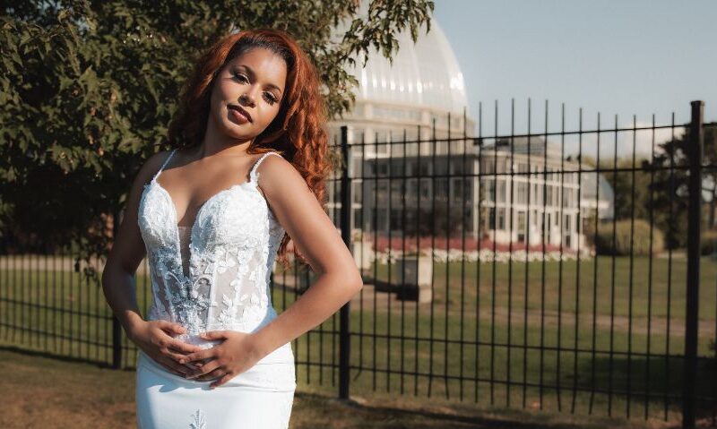 A bride with red hair wears a fitted lace wedding dress while standing outdoors near a glass conservatory behind a black fence.